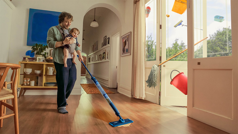 Mężczyzna z dzieckiem na rękach używający mopa elektrycznego Philips OneUp do czyszczenia podłogi