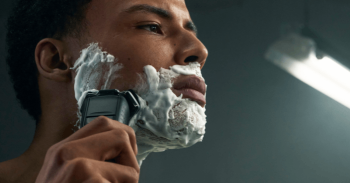 Zbliżenie na mężczyznę o kręconych brązowych włosach, kt&oacute;ry goli sw&oacute;j zarost maszynką elektryczną.