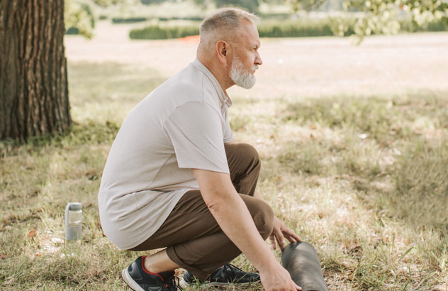Profil mężczyzny z siwymi włosami i białą brodą, kt&oacute;ry zwija matę sportową na łące.