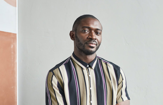 Czarnosk&oacute;ry mężczyzna z bardzo kr&oacute;tko przyciętymi włosami i starannie przyciętą trzydniową brodą patrzy w obiektyw aparatu.