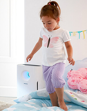 Dziewczynka stojąca na ł&oacute;żku z oczyszczaczem powietrza w tle