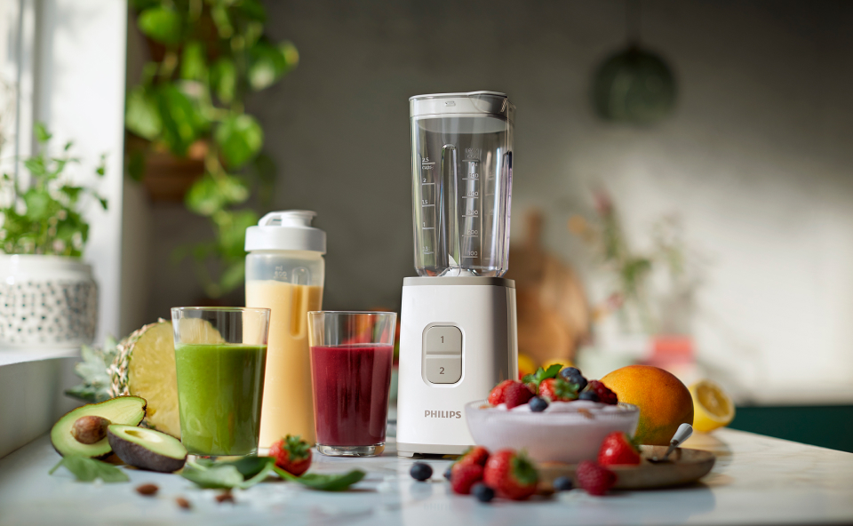 Blender kielichowy stojący na blacie w kuchni, obok są przyrządzone koktajle i leżące owoce.