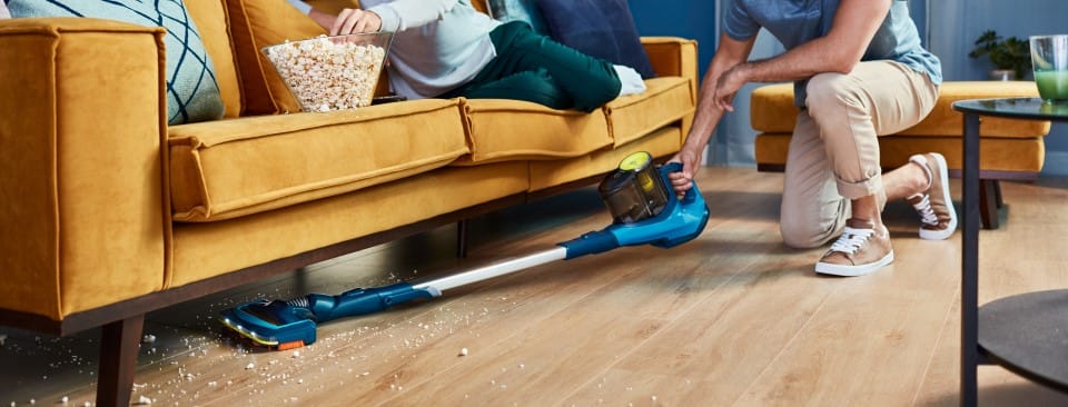 Mężczyzna odkurza przestrzeń pod meblami w wygodny, dokładny i szybki spos&oacute;b dzięki odkurzaczowi bezprzewodowemu.
