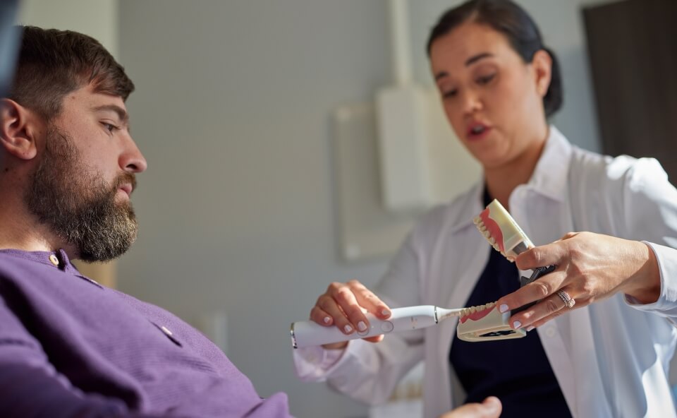 Dentystka w białym uniformie pokazuje mężczyźnie z brodą, jak prawidłowo szczotkować zęby za pomocą elektrycznej szczoteczki i modelu szczęki.