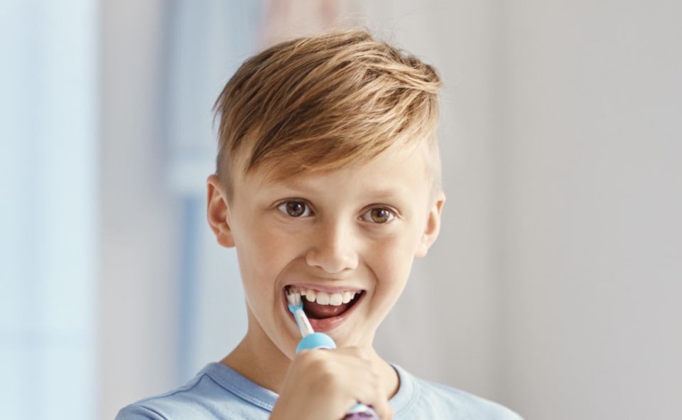 Starszy chłopiec z jasnymi, krótkimi włosami i w jasnoniebieskiej koszulce używa elektrycznej szczoteczki do zębów, szeroko się uśmiechając, stojąc w jasnym, nowoczesnym wnętrzu łazienki.