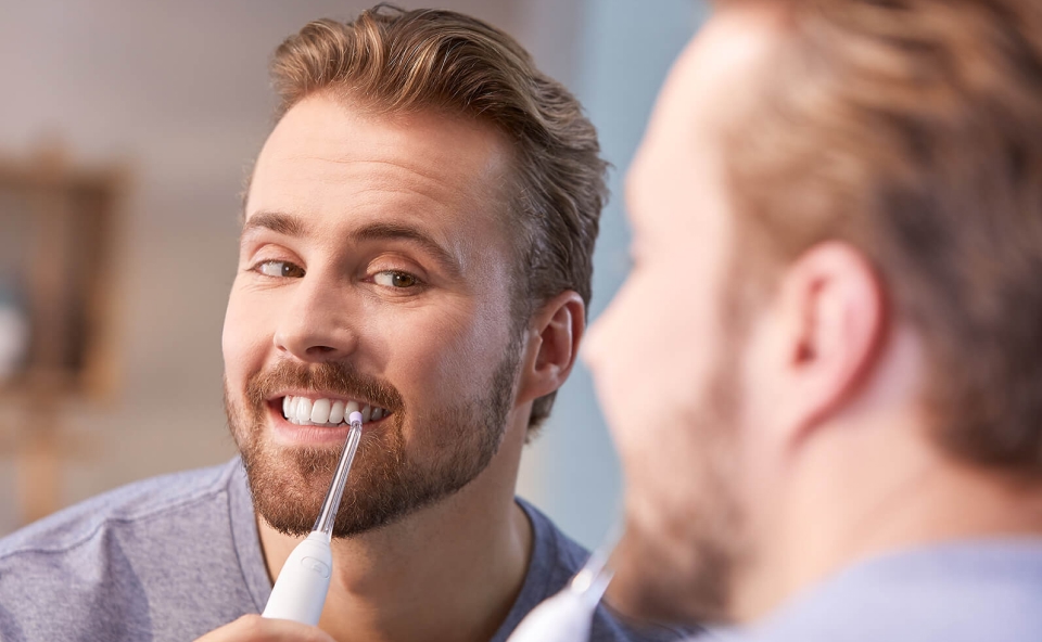 Mężczyzna używający irygatora do zęb&oacute;w, stojący przed lustrem, uśmiecha się, dbając o higienę jamy ustnej.