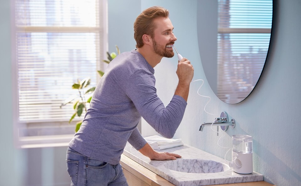 Mężczyzna używający irygatora do zęb&oacute;w przy umywalce, patrząc w lustro, w jasnej łazience z niebieskimi ścianami.