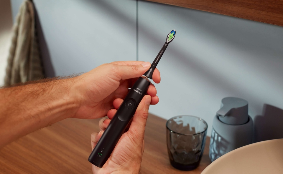 Mężczyzna trzymający w dłoniach czarną szczoteczkę soniczną Philips Sonicare w łazience, na blacie widoczne akcesoria łazienkowe, takie jak szklanka i dozownik mydła.