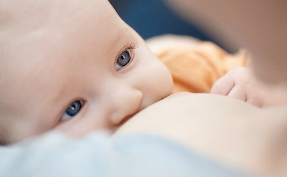 Młoda para leżąca na ł&oacute;żku i bawiąca się z niemowlęciem, kt&oacute;re mężczyzna trzyma nad sobą. Obaj dorośli są szczęśliwi, uśmiechają się szeroko, patrząc na dziecko.