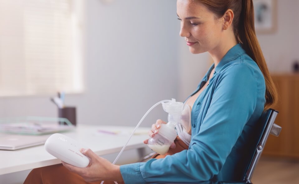 Kobieta używa przenośnego laktatora, siedząc przy biurku, z pojemnikiem na mleko podłączonym do piersi.