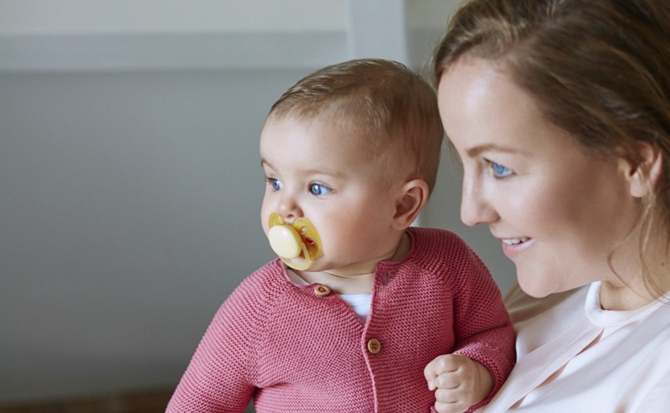 Dziecko ze smoczkiem w buzi trzymane przez mamę na rękach.