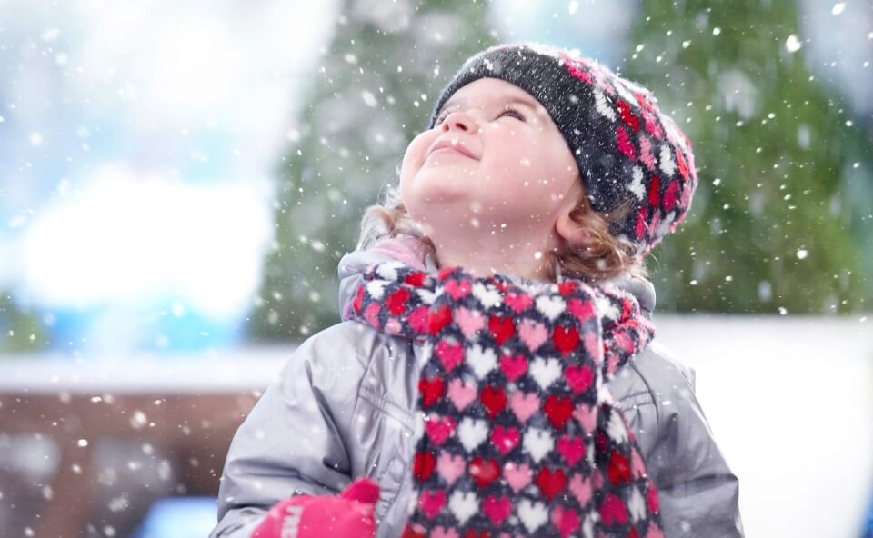 Uśmiechnięte dziecko w zimowym stroju, patrzące w g&oacute;rę podczas opadającego śniegu.