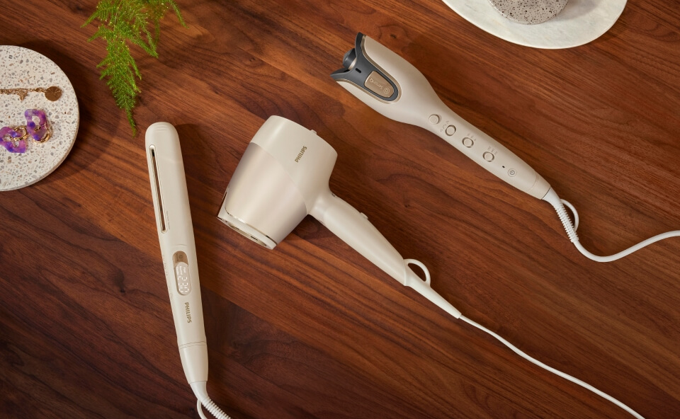 Zestaw beżowych narzędzi do stylizacji włos&oacute;w Philips: prostownica, suszarka i lok&oacute;wka, na drewnianej powierzchni.
