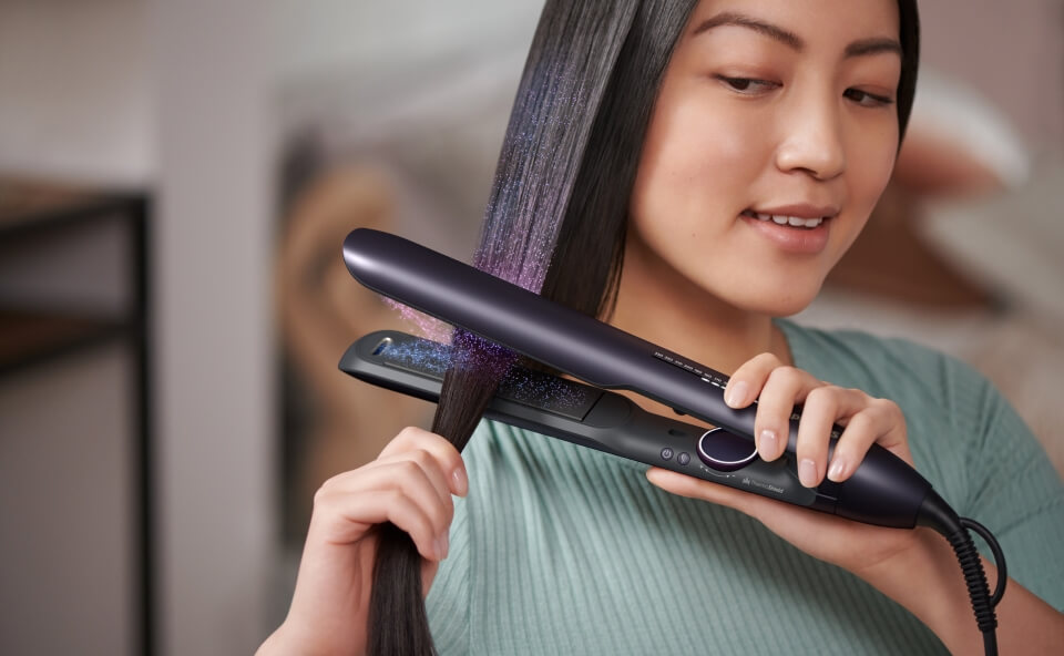 Kobieta używa nowoczesnej prostownicy do stylizacji cienkich włosów, prostując pasmo włosów. Prostownica emituje delikatną mgiełkę, symbolizującą zaawansowane technologie ochronne.