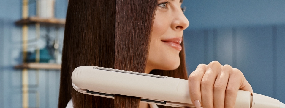 Kobieta prostuje długie, ciemne włosy za pomocą jasnej, nowoczesnej prostownicy. Efekt prostowania sprawia, że włosy są gładkie i lśniące, co sugeruje zastosowanie technologii ochronnych w urządzeniu.
