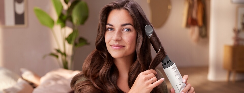 Kobieta z długimi, brązowymi włosami stylizuje je przy użyciu obrotowej suszarko-lokówki. Włosy są delikatnie nawinięte na szczotkę urządzenia. W tle widać jasne wnętrze pokoju z rośliną i wieszakami na ubrania.
